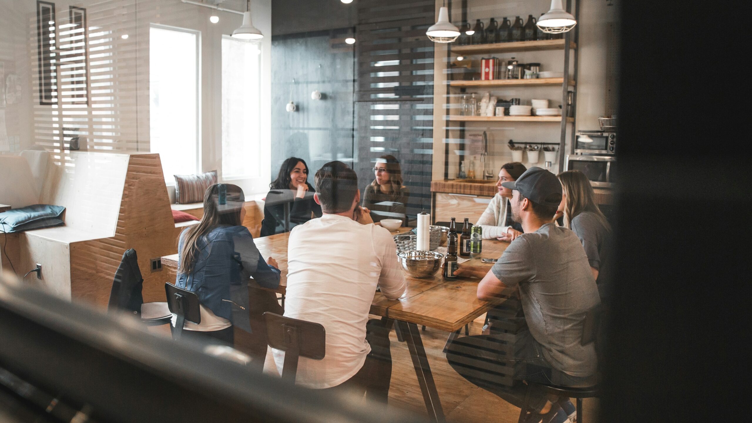 A group of people sitting around a large wooden table in a spacious office kitchen