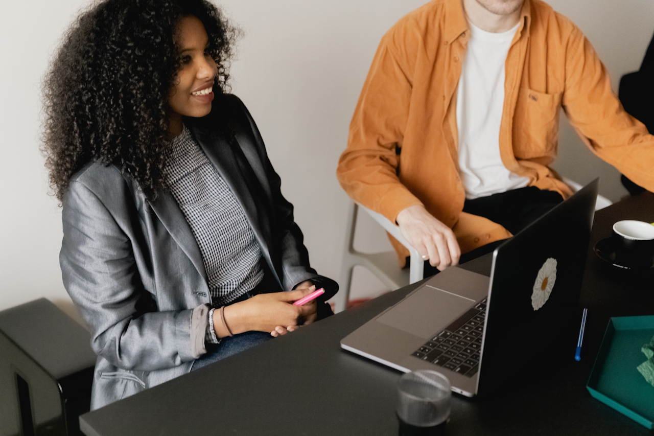 Two people attending a meeting, sit at a desk with a laptop in front of them