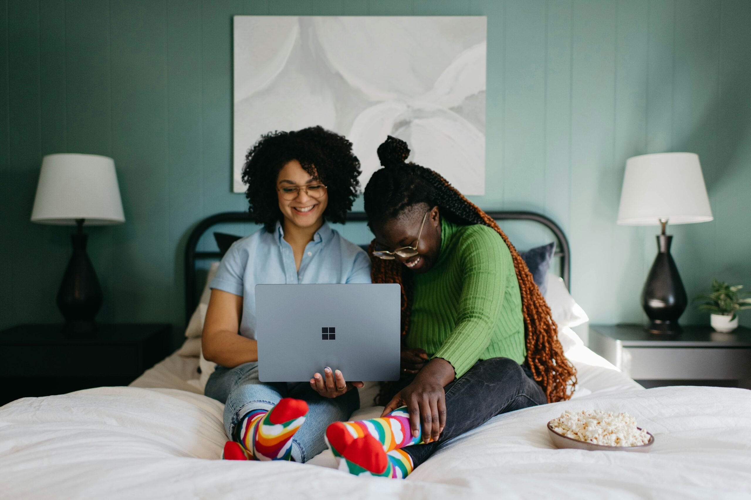 Two smiling women sitting on a bed, looking at the screen of a silver laptop with a bowl of popcorn on the side of the bed
